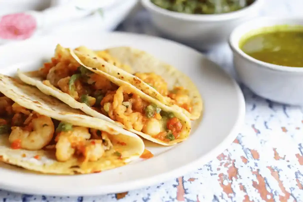 Three tacos gobernador filled with shrimp, cheese, and vegetables on a white plate with green salsa bowls